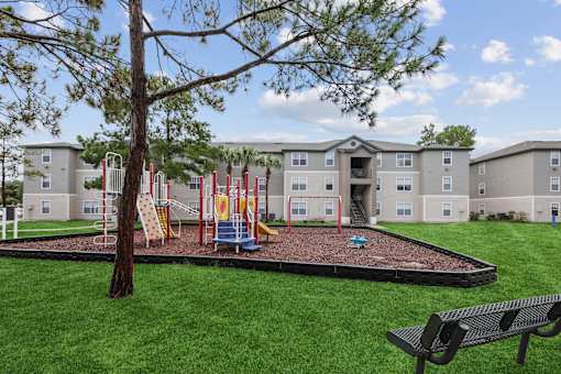 A playground area with a slide, swings, and a tree in front of apartment buildings.