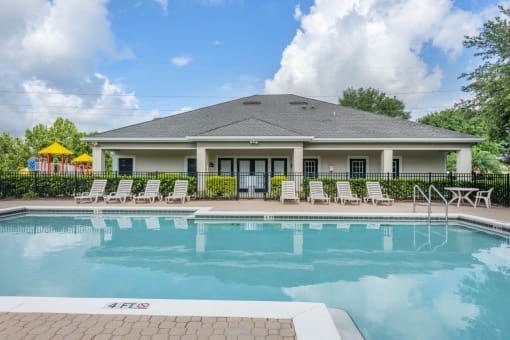 Swimming pool with numerous lounge chairs and a playground and clubhouse in the background.
