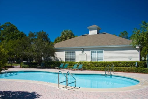 Community pool with sundeck, lounge chairs, surrounded by black metal fence with trees, lake, and trees in the background