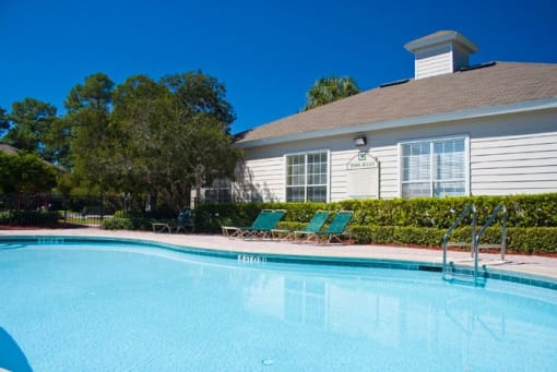 Community pool with sundeck, lounge chairs, surrounded by black metal fence with trees, lake, and trees in the background