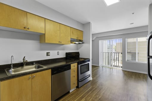 a kitchen with wooden cabinets and stainless steel appliances with a view into the spacious living area with plank flooring and sliding glass doors at Illumina Apartment Homes, Seattle, WA