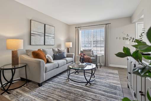 A living room with a grey couch, a glass coffee table, and a large window with blinds.