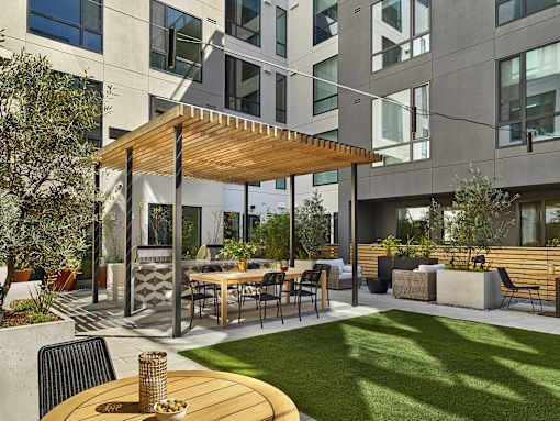 A patio with a table and chairs is surrounded by a grassy area and a building with large windows.