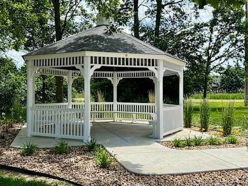 A white gazebo with a black roof is surrounded by gravel and greenery.