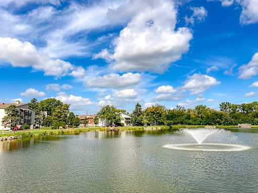 A fountain in the middle of a lake with buildings and trees in the background.