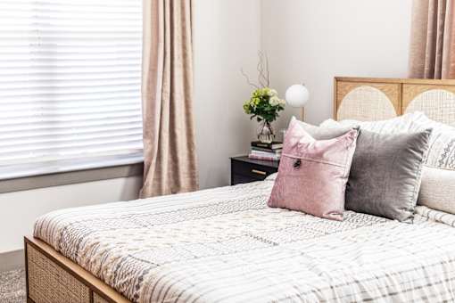 A bed with a white comforter and a pink pillow is in the foreground of a room with a window and curtains.