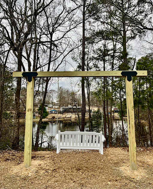 A white bench is situated under a wooden structure in a wooded area.