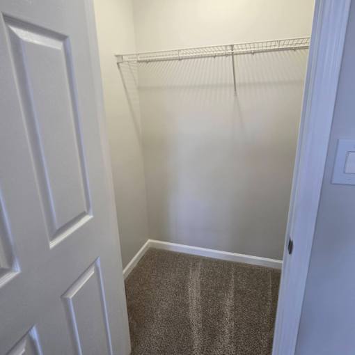 A white door is on the left of a hallway with a carpeted floor and a wire shelf on the wall.