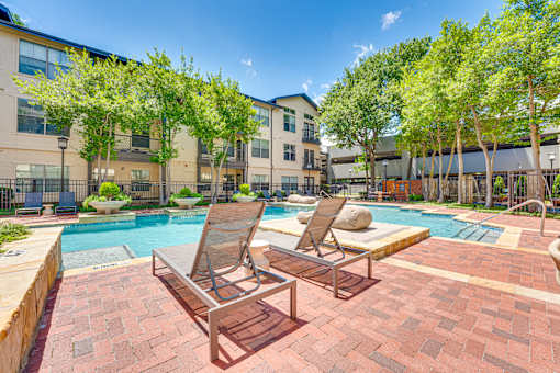 Two lounge chairs sit on a brick patio next to a pool.