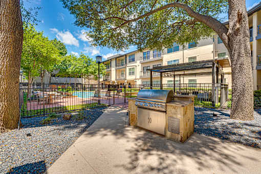 A BBQ grill is in the middle of a sandy area with a building in the background.