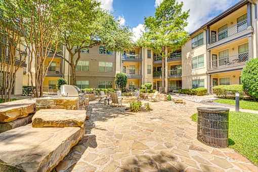 A stone walkway leads through a landscaped courtyard with a trash can and trees.