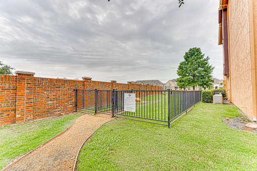A black metal fence surrounds a grassy area with a brick wall on one side and a dirt path leading to a gate at Limestone Ranch Apartments, Lewisville 75067