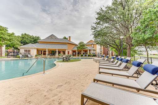 A row of lounge chairs are set up on a sandy area in front of a pool and a building at Limestone Ranch Apartments, Lewisville, Texas
