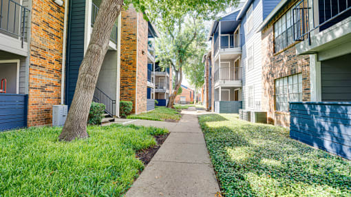a sidewalk between two apartment buildings with grass and trees at The Manhattan Apartments, Dallas, TX