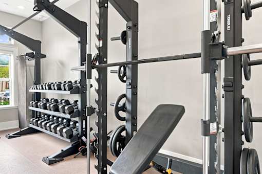 A black weight bench is in front of a squat rack with dumbbells on the rack.