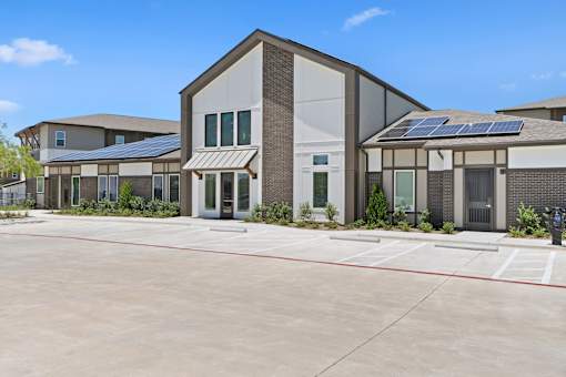 Exterior view of the community clubhouse building featuring modern architecture, solar panels, landscaped grounds, resident parking spaces, and a main entrance with decorative windows.