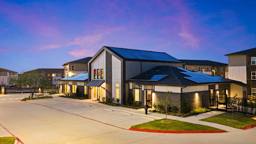 Exterior view of the community clubhouse building at dusk, featuring modern architecture, landscaped grounds, solar panels, illuminated windows, and adjacent resident parking.