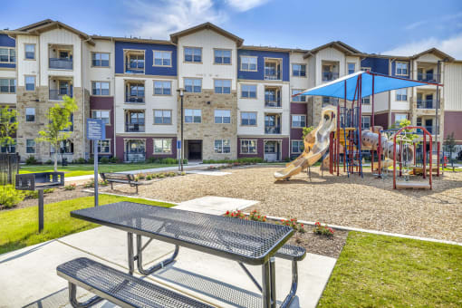 an outdoor playground with a slide and picnic table in front of an apartment building
