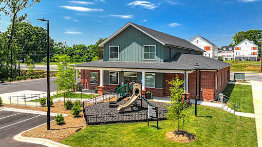 Exterior view of a community clubhouse and playground area, featuring a brick and siding building with a covered porch, fenced play structure with slides, and landscaped grounds.