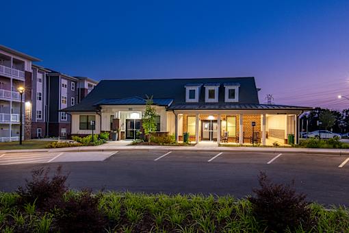 Residential clubhouse exterior with a wraparound covered entry, neutral siding, and landscaped surroundings adjacent to a paved parking lot.