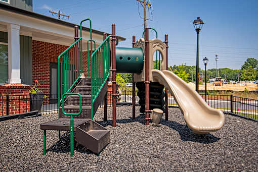 Children’s playground featuring a slide, climbing tunnel, and stair access, positioned within a fenced space beside a residential clubhouse.