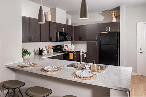 Modern apartment kitchen with dark cabinetry, black appliances, and pendant lighting, featuring an island countertop with double sink and bar seating.