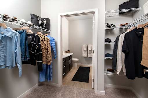 Spacious walk-in closet with built-in wire shelving and hanging space on both sides, offering ample storage for clothing, shoes, and accessories, with direct access to a modern en-suite bathroom featuring a large vanity and updated finishes.