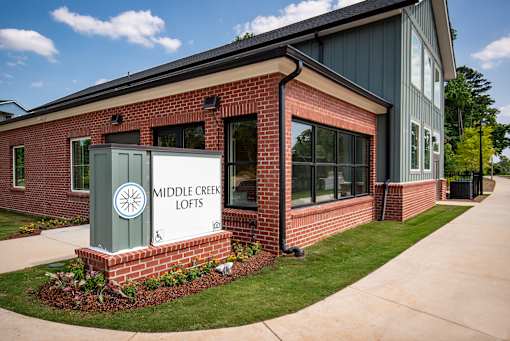 Apartment community sign mounted on a brick base with surrounding landscaping, positioned next to a clubhouse building with siding and large windows.