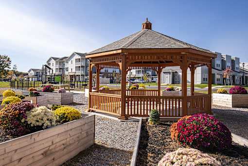A wooden gazebo is surrounded by a gravel area and flower beds.