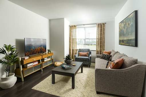 A living room with a grey couch, a wooden coffee table, a painting on the wall, and a large window with curtains.