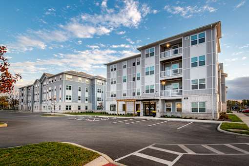 Exterior view of The Lofton, a modern four-story apartment building in Columbus, OH with balconies, parking lot, and landscaped entry.