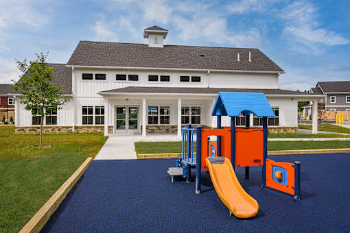 A playground with a yellow slide and orange and blue play structures in front of a white building with a black roof.