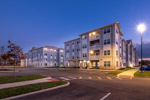 Evening view of apartment community with modern multi-story buildings, illuminated entrances, and surrounding parking areas under a twilight sky.