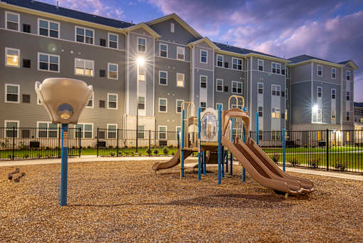 Community playground with slides and climbing structures set within a landscaped outdoor area, with apartment buildings in the background.
