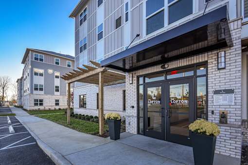 Exterior view of apartment community entrance featuring glass double doors, modern brick façade, and covered walkway leading to the leasing office.