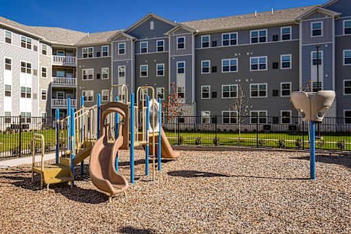 Community playground with slides and climbing structures set within a landscaped outdoor area, with apartment buildings in the background.