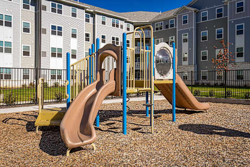 Community playground with slides and climbing structures set within a landscaped outdoor area, with apartment buildings in the background.