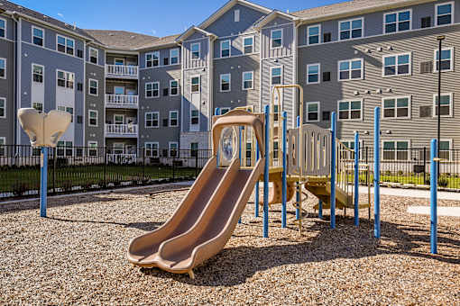 Community playground with slides and climbing structures set within a landscaped outdoor area, with apartment buildings in the background.