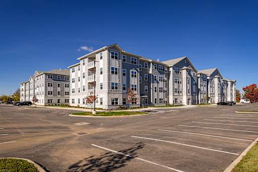 Exterior view of modern apartment buildings with clean architectural lines, surrounding parking area, and landscaped grounds under a clear blue sky.