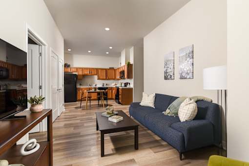 Model apartment living room and kitchen at The Davis II in Cleveland, featuring a sofa, coffee table, wood-style flooring, and an open-concept kitchen with wood cabinetry and black appliances.