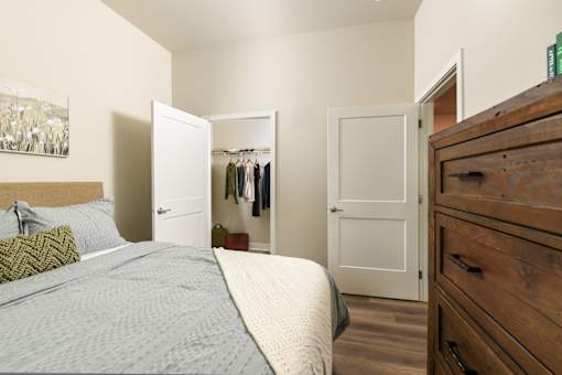 Model apartment bedroom at The Davis II in Cleveland, showing a bed beside an open closet with hanging clothes, wood-style flooring, and an open doorway leading to the hallway.