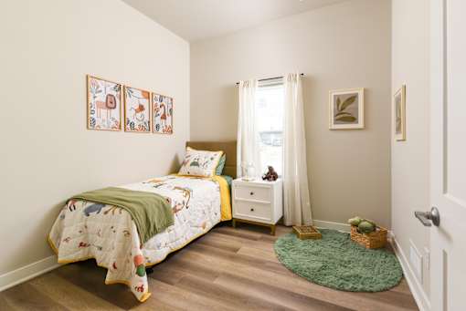 Model apartment bedroom at The Davis II in Cleveland, featuring a twin bed with neutral bedding, decorative pillows, a bedside table, wood-style flooring, and a window with natural light.