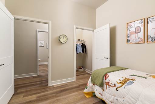 Model apartment bedroom at The Davis II in Cleveland, featuring a furnished bed, wood-style flooring, neutral walls, wall décor, and an open doorway leading to the hallway.