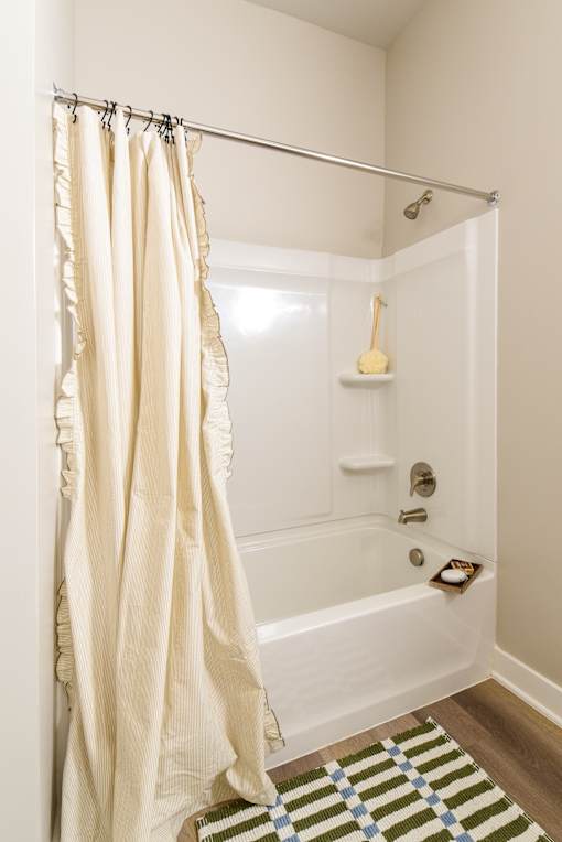 Model apartment bathroom at The Davis II in Cleveland, featuring a white bathtub and shower combination with a curtain, built-in shelving, and chrome fixtures.