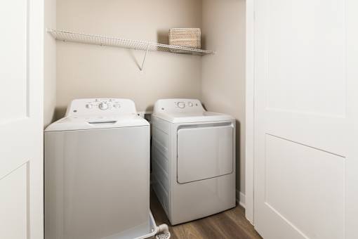 Model apartment laundry area at The Davis II in Cleveland, featuring a white top-loading washing machine and front-loading dryer with wall-mounted shelving above.