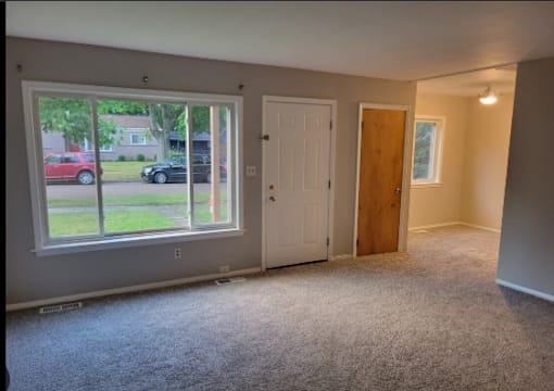 A room with a carpeted floor, a white door, a window with a view of a street, and a wooden door.