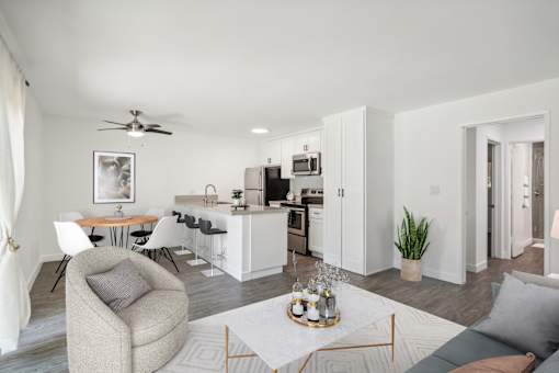 A modern living room with a grey sofa, a grey armchair, a white coffee table, a white dining table, a white kitchen, a white ceiling fan, a white rug, a white wall, a white door, a