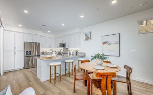 A kitchen with a table and chairs in the foreground and a fridge and microwave in the background.