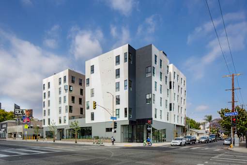 A modern building with a white and grey exterior is situated on a street corner with cars and traffic lights.