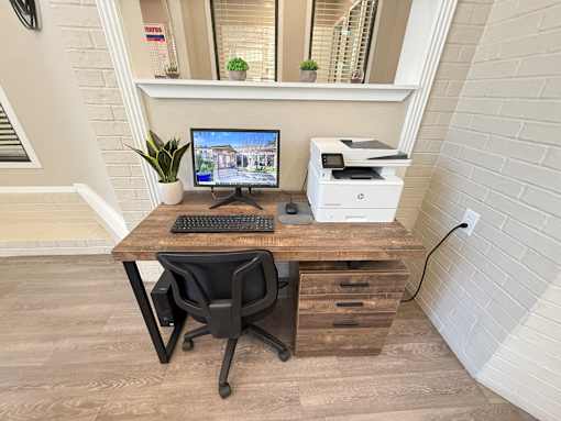 A computer desk with a monitor, keyboard, and printer on a wooden desk with a black chair.
